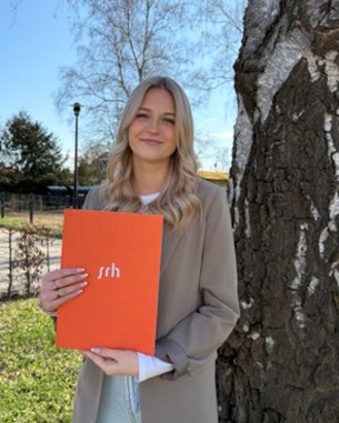 Eine blonde Frau hält eine orangefarbene Mappe mit der Aufschrift "srh" vor einem Baum im Freien.