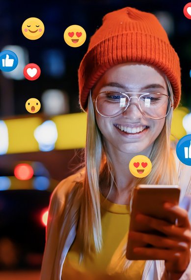 Eine lächelnde Frau mit Brille und rotem Hut schaut auf ihr Smartphone, umgeben von Symbolen für Likes und Emojis, während sie in einer städtischen Umgebung steht.
