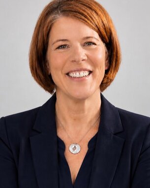 Eine lächelnde Frau mit schulterlangen, roten Haaren trägt einen dunkelblauen Blazer und eine Halskette. Sie hat die Arme verschränkt.