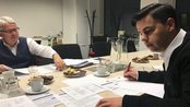Zwei Männer sitzen an einem Tisch, der mit Dokumenten, Kaffeetassen und Gebäck gedeckt ist. Der Mann links trägt eine Weste und schaut nachdenklich, während der Mann rechts Notizen macht und einen Stift hält.