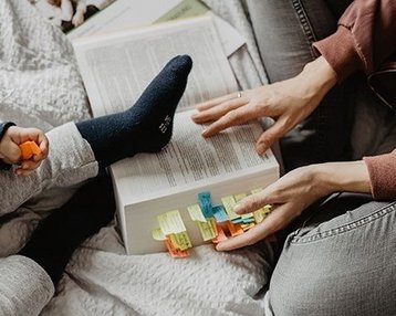 Eine Hand hält ein Buch mit bunten Markierungen, während ein Kind mit einem Fuß auf dem Buch sitzt. Die andere Hand des Erwachsenen zeigt auf die Markierungen.