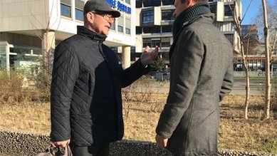Zwei Männer stehen im Freien und führen ein Gespräch. Der Mann links trägt eine dunkle Jacke und eine Mütze, während der Mann rechts einen grauen Mantel und einen Schal trägt.