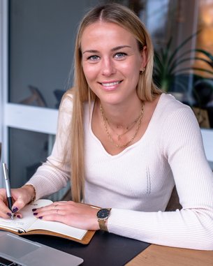 Eine junge Frau sitzt an einem Tisch, schreibt in ein Notizbuch und schaut lächelnd in die Kamera. Ein Laptop und eine Pflanze sind sichtbar.