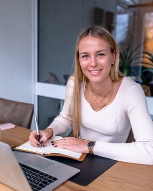 Eine junge Frau sitzt an einem Tisch, schreibt in ein Notizbuch und schaut lächelnd in die Kamera. Ein Laptop und eine Pflanze sind sichtbar.