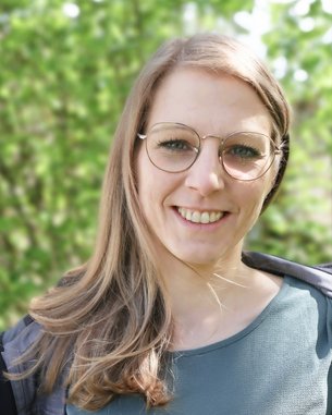 Eine lächelnde Frau mit langen, blonden Haaren und Brille steht vor einem unscharfen, grünen Hintergrund. Sie trägt ein graues Oberteil und hat eine freundliche Ausstrahlung.