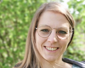 Eine lächelnde Frau mit langen, blonden Haaren und Brille steht vor einem unscharfen, grünen Hintergrund. Sie trägt ein graues Oberteil und hat eine freundliche Ausstrahlung.