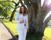 Eine Frau in einem weißen Anzug steht neben einem großen Baum und hält ein Notizbuch. Die Umgebung ist sonnig und grün.