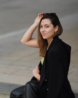 Eine Person sitzt auf einer Treppe, trägt einen schwarzen Blazer, ein gelbes Oberteil und schwarze Hosen. Sie hat lange Haare und lächelt.