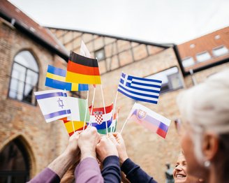Mehrere Hände halten verschiedene Nationalflaggen, darunter Deutschland, Griechenland, Israel, Kroatien und Russland, in einer Feierlichkeit im Freien.