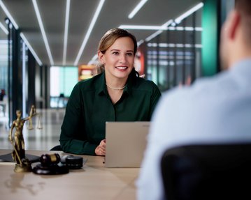 Eine Frau in einem grünen Hemd sitzt an einem Tisch und spricht mit einem Mann. Auf dem Tisch steht eine Statue der Justitia.