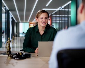 Eine Frau in einem grünen Hemd sitzt an einem Tisch und spricht mit einem Mann. Auf dem Tisch steht eine Statue der Justitia.