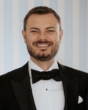 Ein Mann mit Bart trägt einen schwarzen Anzug und eine Fliege. Er lächelt freundlich in die Kamera, vor einem Hintergrund mit blauen Streifen.