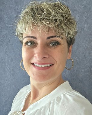 Frau mit kurzem, lockigem Haar und großen Ohrringen, die in die Kamera lächelt. Sie trägt ein helles Oberteil und hat einen neutralen Hintergrund.