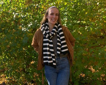 Eine Person steht in einem herbstlichen Park, trägt eine braune Jacke und einen schwarz-weißen gestreiften Schal. Im Hintergrund sind grüne Sträucher und gelbe Blätter zu sehen.