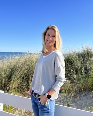 Eine Frau mit langen, blonden Haaren steht an einem Strand, umgeben von Gras und einem weißen Zaun. Sie trägt einen grauen Pullover und Jeans, während sie in die Kamera lächelt.