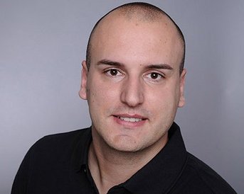 Ein Mann mit kurzem, glattem Haar trägt ein schwarzes Polohemd und blickt direkt in die Kamera. Der Hintergrund ist neutral und grau.