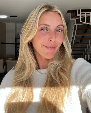 Eine Person mit langen, blonden Haaren lächelt in die Kamera, während sie in einem hellen Raum mit moderner Einrichtung steht.