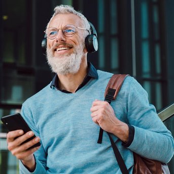 Ein älterer Mann mit Brille und Kopfhörern lächelt, während er ein Smartphone in der Hand hält. Er trägt einen hellblauen Pullover und eine braune Tasche.