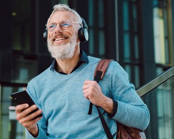 Ein älterer Mann mit Brille und Kopfhörern lächelt, während er ein Smartphone in der Hand hält. Er trägt einen hellblauen Pullover und eine braune Tasche.