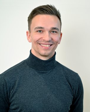 Ein junger Mann mit kurzen, gestylten Haaren trägt einen schwarzen Rollkragenpullover und lächelt freundlich in die Kamera. Der Hintergrund ist neutral und hell.