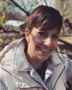 Eine Frau mit kurzem, braunem Haar trägt eine graue Jacke und lächelt freundlich. Im Hintergrund sind unscharfe Bäume und ein Gebäude zu sehen.