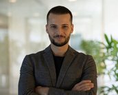 Ein Mann mit kurzem Bart und kurzen Haaren steht mit verschränkten Armen in einem modernen Raum, umgeben von Pflanzen. Er trägt einen grauen Blazer und blickt direkt in die Kamera.