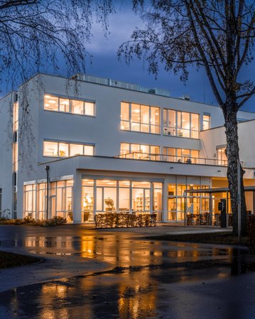 Moderne, mehrstöckige Gebäude mit großen Fenstern, beleuchtet bei Dämmerung. Nasser Parkplatz im Vordergrund spiegelt die Lichter wider.