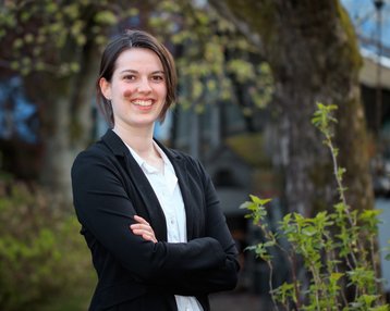 Eine Person mit kurzen Haaren trägt einen schwarzen Blazer und steht mit verschränkten Armen in einer grünen Umgebung. Sie lächelt freundlich in die Kamera.