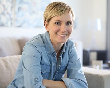 Psychologie | SRH Fernhochschule Eine lächelnde Frau mit kurzen blonden Haaren sitzt auf einem Sofa in einem hellen Raum. Sie trägt ein blaues Jeanshemd und blickt freundlich in die Kamera.