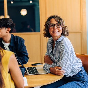 Eine lächelnde Frau mit lockigem Haar und Brille sitzt an einem Tisch mit einem Laptop, umgeben von anderen Personen in einem modernen Raum.