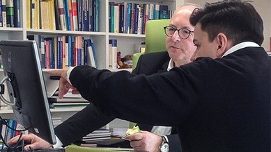 Zwei Personen sitzen an einem Schreibtisch, einer zeigt auf den Computerbildschirm, während der andere aufmerksam zuschaut und ein Stück Obst hält. Im Hintergrund sind Bücherregale sichtbar.