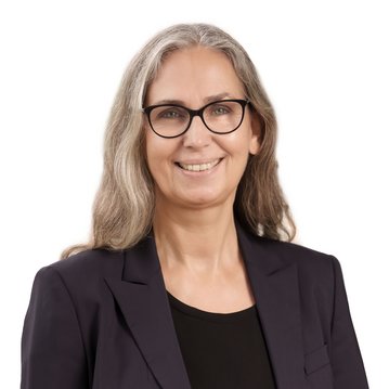 Eine lächelnde Frau mit langen, grauen Haaren und Brille trägt einen schwarzen Blazer. Sie steht vor einem neutralen Hintergrund.