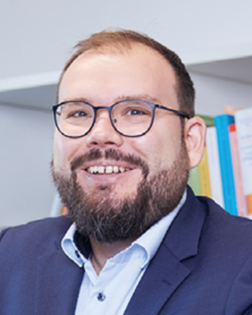 Ein Mann mit Brille und Bart lächelt in einem Büro, trägt einen Anzug und sitzt vor einem Bücherregal.