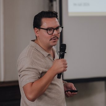 Ein Mann mit Brille hält ein Mikrofon und spricht vor einer Präsentationsleinwand. Er trägt ein helles, kurzärmliges Hemd und hat dunkle, kurze Haare.