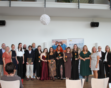 Gruppe von Menschen in festlicher Kleidung, die auf einer Veranstaltung zusammenstehen, einige halten Blumen. Im Hintergrund ein Banner mit dem Text „DER PFLEGEMANAGER“.