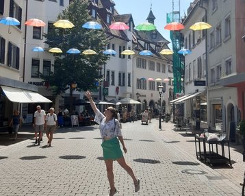 Eine Frau in einem grünen Rock und einem weißen Oberteil steht in einer belebten Straße, während bunte Regenschirme über ihr hängen. Die Umgebung zeigt historische Gebäude und Passanten, die die Straße entlanggehen.