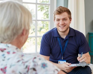 Ein junger Mann in einem blauen Poloshirt spricht lächelnd mit einer älteren Frau, während er Notizen auf einem Notizblock macht.