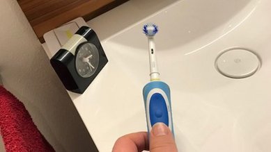 Eine Hand hält eine elektrische Zahnbürste über einem Waschbecken. Im Hintergrund ist eine Wanduhr und ein rotes Handtuch sichtbar.