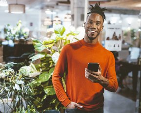 Ein Mann mit einem breiten Lächeln trägt einen orangefarbenen Pullover und hält ein Smartphone in der Hand, umgeben von grünen Pflanzen in einem modernen Innenraum.