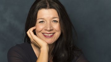 Eine Frau mit langen, dunklen Haaren lächelt freundlich, während sie ihren Kopf leicht zur Seite neigt und eine Hand an ihr Gesicht legt. Der Hintergrund ist einfarbig und neutral.