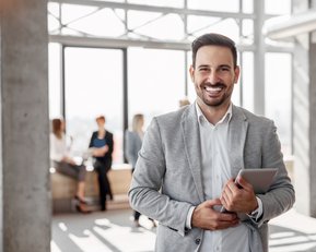 Ein lächelnder Mann in einem grauen Anzug hält ein Tablet in der Hand. Im Hintergrund sind mehrere Personen in einem modernen Büro zu sehen, die miteinander sprechen.