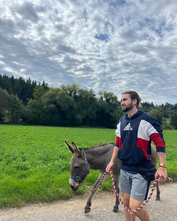 Ein Mann führt einen Esel an der Leine auf einem Weg entlang einer grünen Wiese unter einem bewölkten Himmel.
