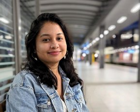Eine junge Frau mit langen, dunklen Haaren sitzt auf einer Bank, trägt eine Jeansjacke und lächelt in einem modernen Innenraum.