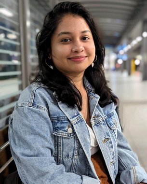 Eine junge Frau mit langen, dunklen Haaren sitzt auf einer Bank, trägt eine Jeansjacke und lächelt in einem modernen Innenraum.