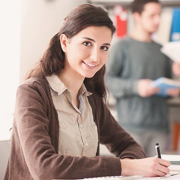 Eine junge Frau sitzt an einem Tisch und schreibt, während zwei Personen im Hintergrund stehen und Unterlagen halten. Der Tisch ist mit Schreibutensilien und einem Notizbuch ausgestattet.