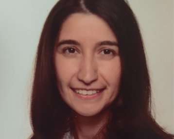 Eine Frau mit langen, dunklen Haaren und einem freundlichen Lächeln trägt ein helles Hemd. Der Hintergrund ist neutral und hell.