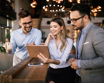 Drei Personen in einem modernen Café betrachten gemeinsam ein Tablet. Zwei Männer, einer mit Brille, halten eine Tasse, während die Frau lächelt und auf den Bildschirm zeigt.