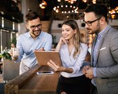 Drei Personen in einem modernen Café betrachten gemeinsam ein Tablet. Zwei Männer, einer mit Brille, halten eine Tasse, während die Frau lächelt und auf den Bildschirm zeigt.