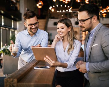 Drei Personen in einem modernen Café betrachten gemeinsam ein Tablet. Zwei Männer, einer mit Brille, halten eine Tasse, während die Frau lächelt und auf den Bildschirm zeigt.