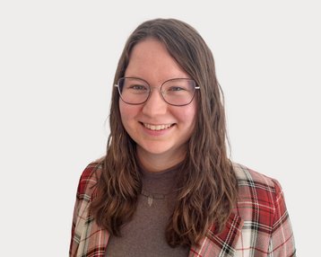 Eine lächelnde Person mit langen, lockigen Haaren trägt eine kariert gestreifte Jacke und eine Brille. Der Hintergrund ist neutral.